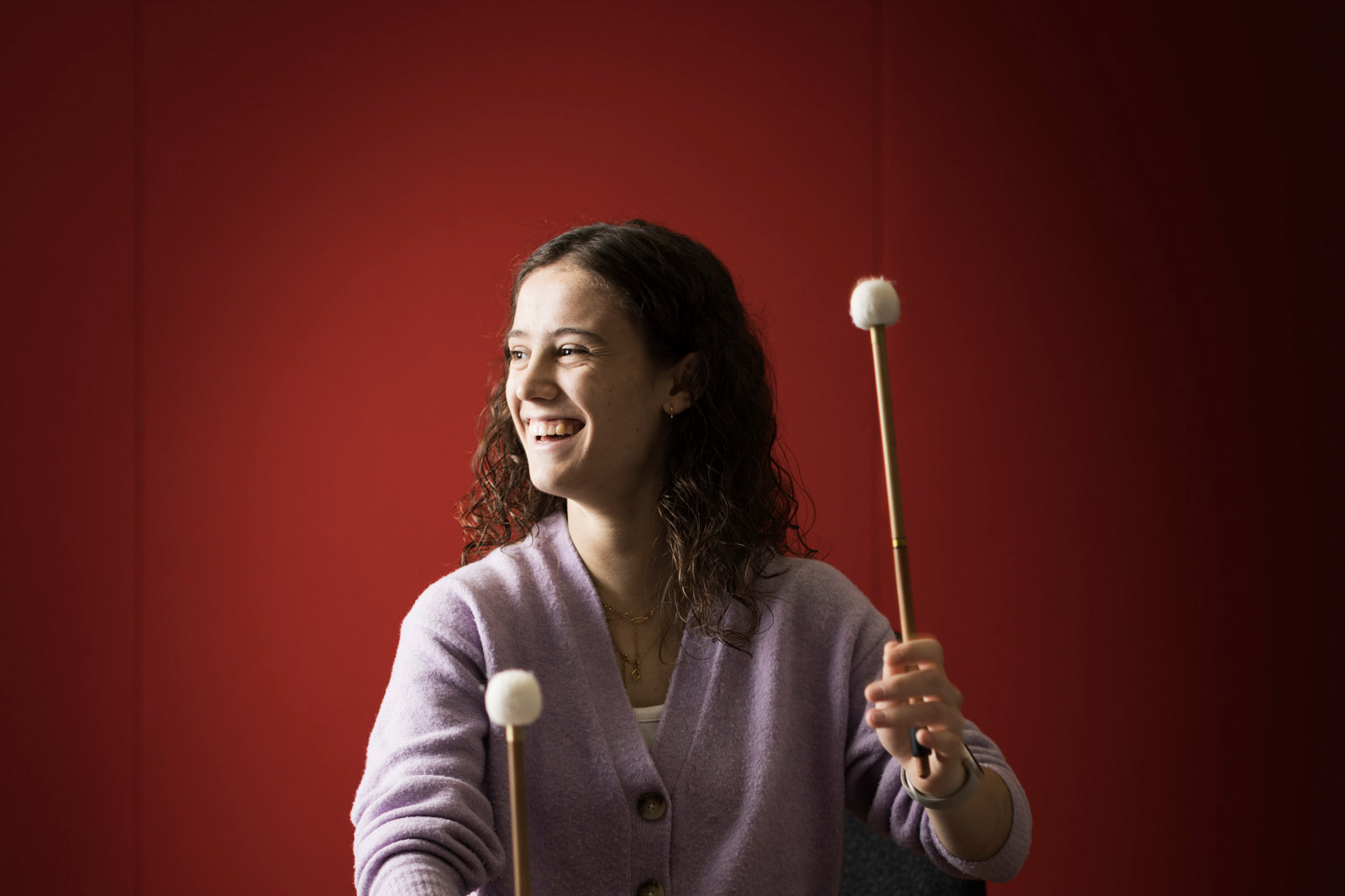 A women playing the timpani and looking to the side, with a deep, rose pink background.